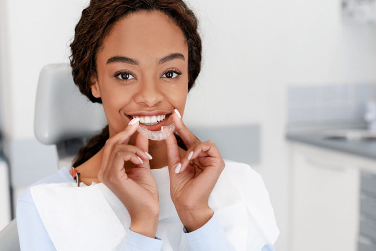 An African American woman sits in a dental chair and holds an Invisalign tray to her mouth