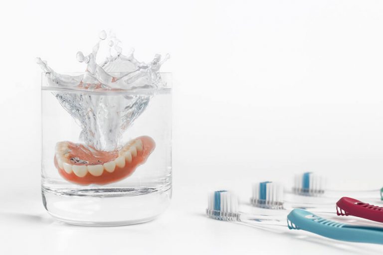 Dentures being dropped into a glass of water with a row of toothbrushes to the right of the glass